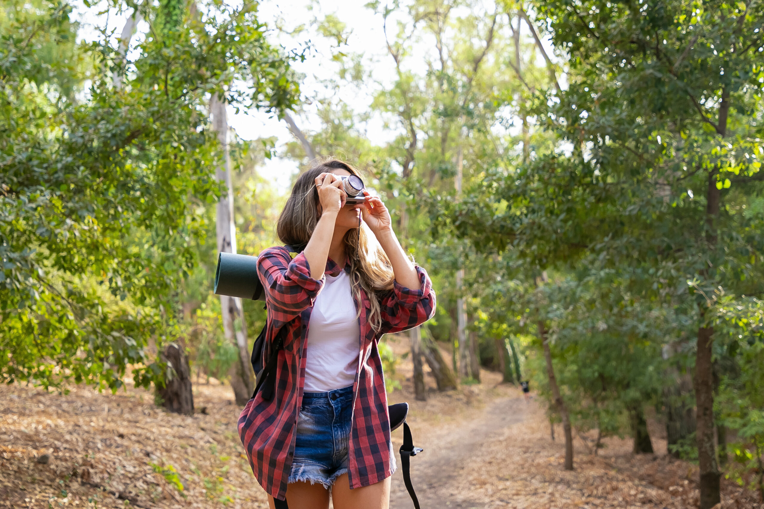 Long-haired woman taking photo of nature and standing on road in forest. Blonde Caucasian lady holding camera and shooting landshate. Backpacking tourism, adventure and summer vacation concept